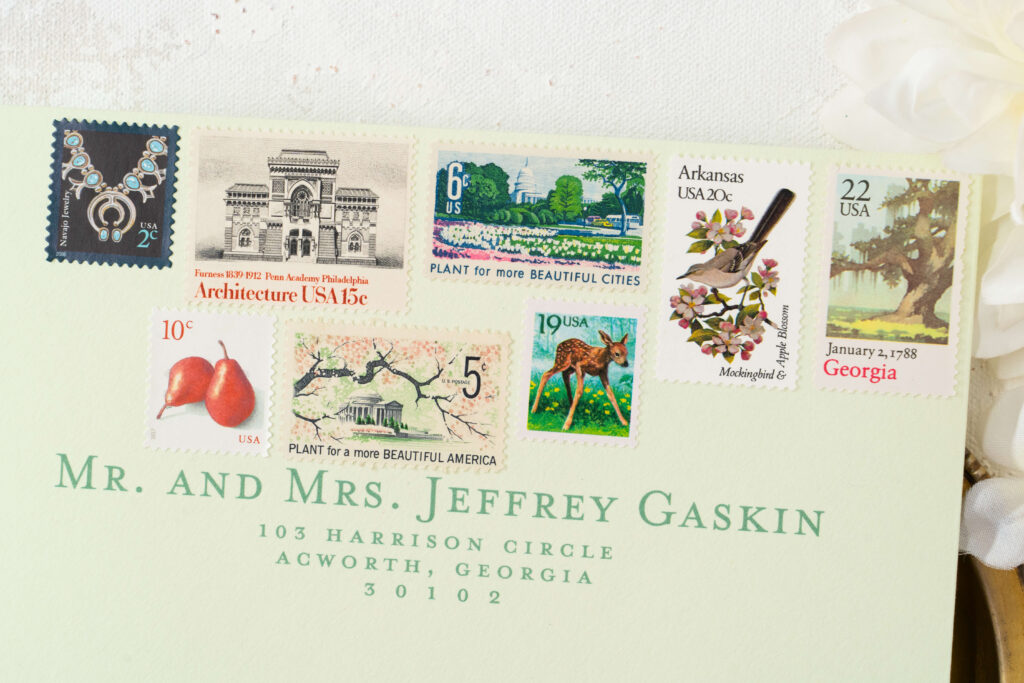 Eight vintage stamps sit beautifully on top of a light green envelope. A Georgia vintage stamp, a state flower and state bird stamp, a Plant for More Beautiful Cities vintage stamp, an architecture vintage stamp, a 2¢ stamp featuring Navajo jewelry, a 10¢ pear stamp, a Plant for a more Beautiful America stamp, and a whimsical fawn vintage stamp.