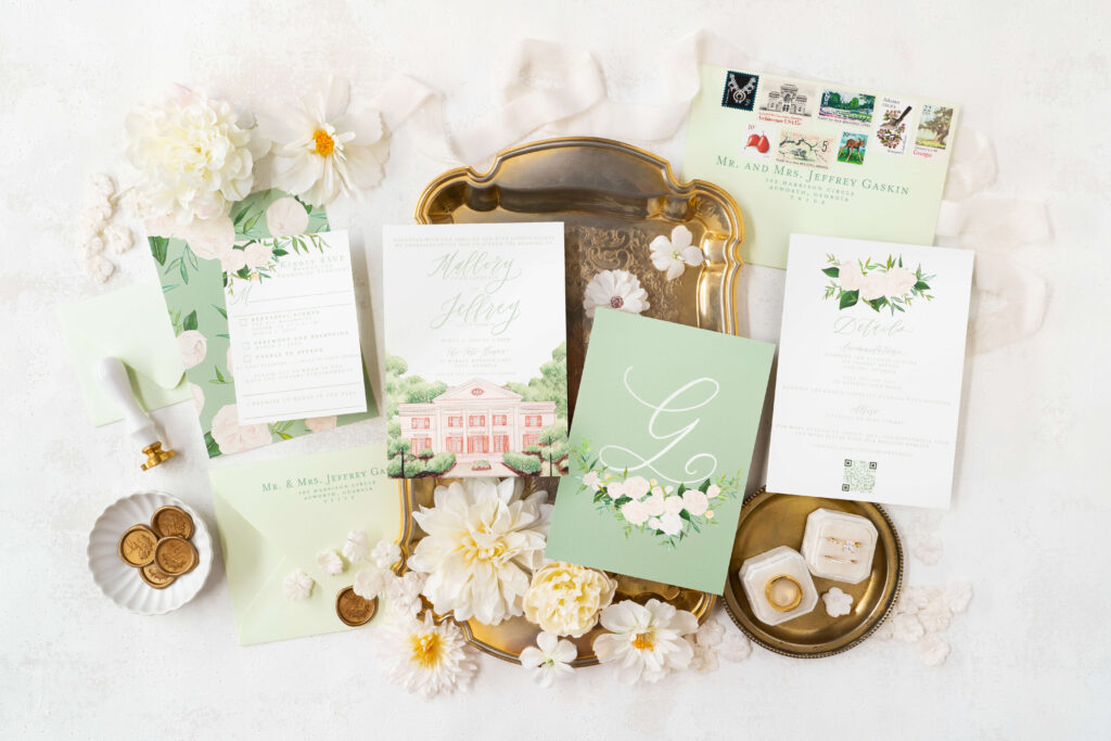 Elegant flat lay of a custom wedding invitation suite designed for Mallory and Jeff's fairytale wedding at Tate House. The suite features a beautifully illustrated invitation with a watercolor painting of the historic Tate House, complemented by sage green envelopes adorned with vintage stamps. Hand-lettered calligraphy adds a personal touch to each piece, including the details card and RSVP card. The arrangement is enhanced with delicate white florals, a golden tray, wax seals, and wedding rings, showcasing a perfect blend of timeless romance and sophisticated charm.