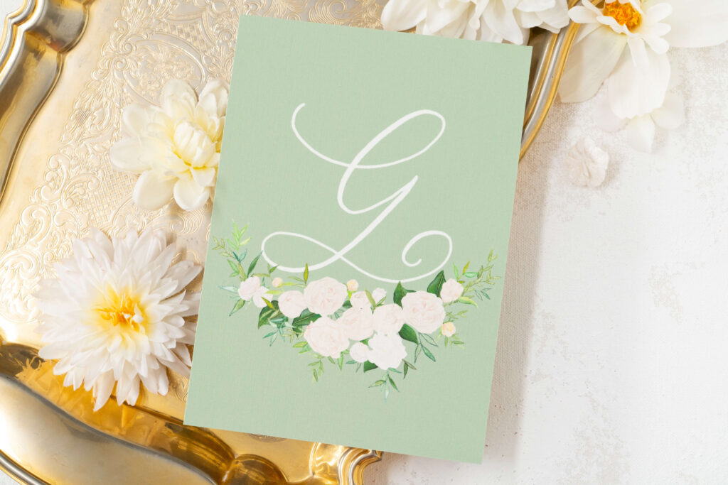 The back of Mallory's formal invitation features their custom wedding crest in watercolor. This acts as a beautiful monogram in hand-lettered calligraphy on a sage card with white watercolor florals below a calligraphy capital G.