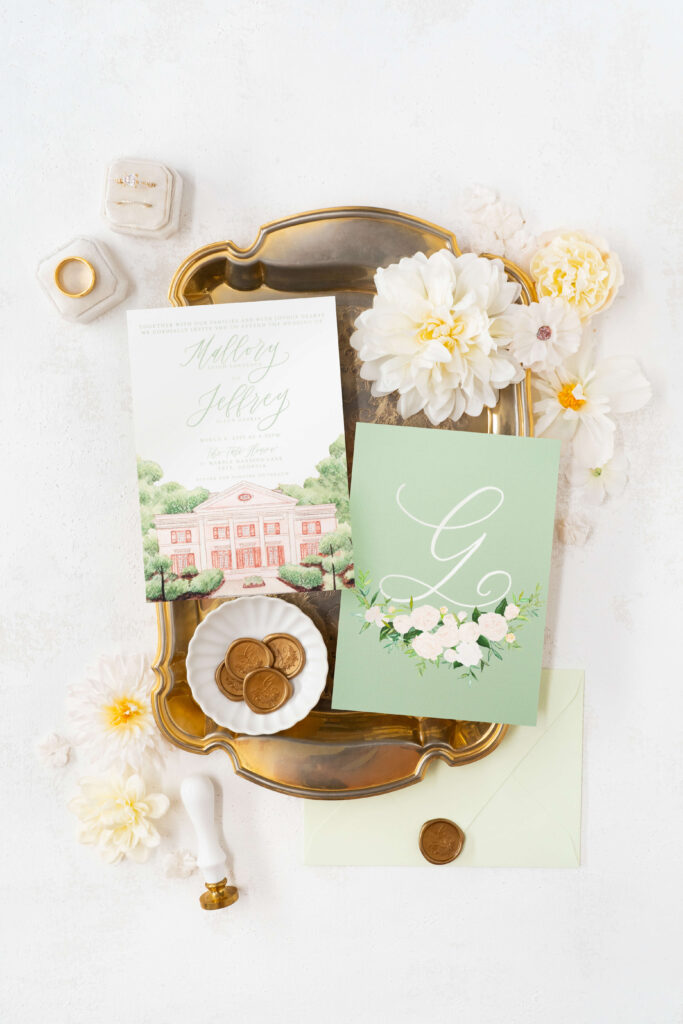 Sophisticated flat lay of sage green wedding invitations. Custom gold wax seals and gold accents are showcase alongside the formal invitation front and back, showing watercolor artwork of the Tate House in Tate, Georgia on the front, and a custom monogram with white watercolor roses on the back.