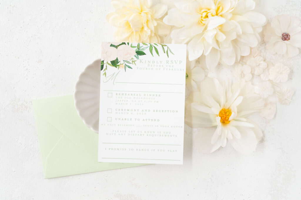 Custom RSVP card surrounded by white flowers with a light green RSVP envelope. The RSVP card has watercolor white roses and greenery in the top corner and space for guests to write their names and check which events they are able to attend.