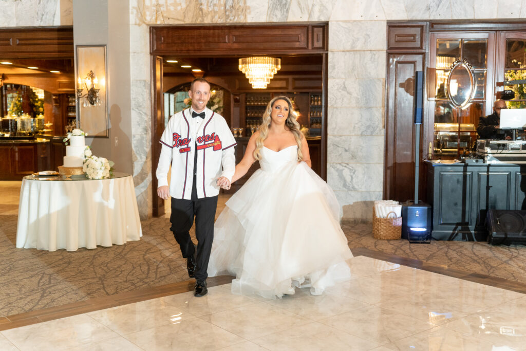 Bride and Groom enter wedding reception hand in hand. Groom is wearing an Atlanta Braves baseball jersey over his suit, showing his love for MLB baseball at his wedding.