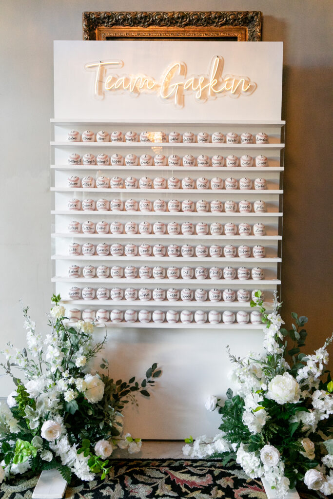 Wedding seating chart display with baseballs, each featuring guest names and table numbers in hand-lettered modern calligraphy. The white shelving unit is adorned with a neon sign reading 'Team Gaskin' at the top, and flanked by lush white floral arrangements, blending the couple's love for baseball with elegant wedding decor.