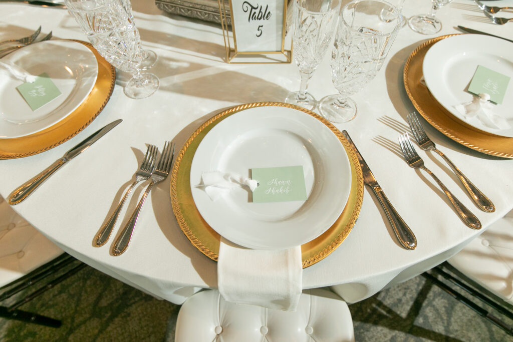 Elegant and modern white and gold place setting with a single sage green place card with white silk ribbon on the plate. The place card has guest's name hand-lettered in white calligraphy.