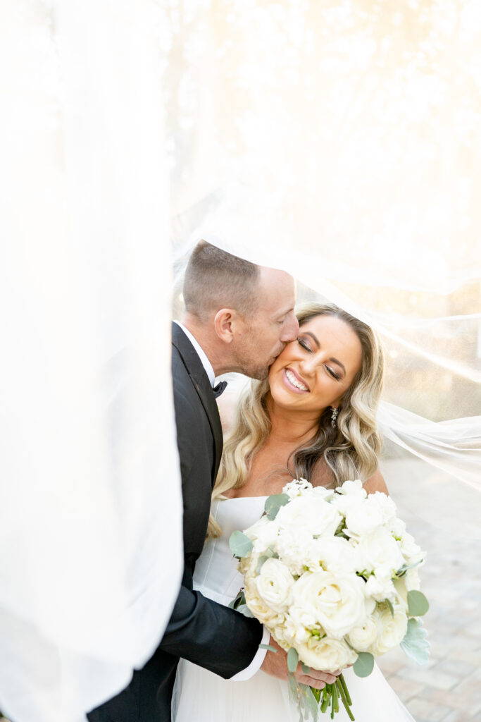 Mallory and Jeff, E Stokes Creations clients for wedding stationery and custom day-of goods share a kiss under her veil on their wedding day. Mallory is holding a bouquet of white roses, peonies, and ranunculus.