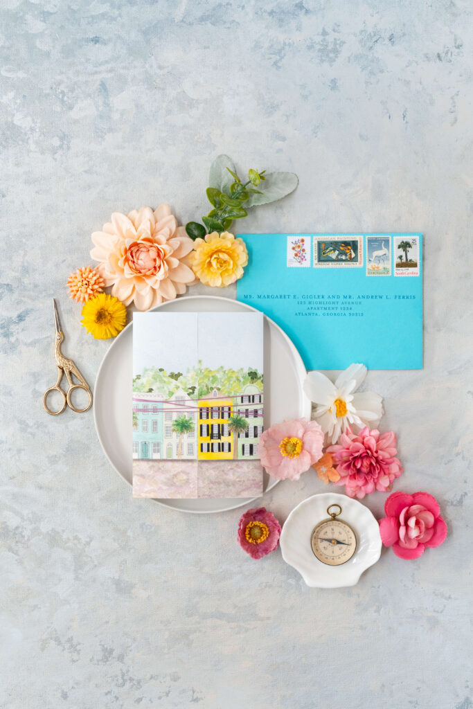 Flatlay of watercolor gatefold card showcasing Rainbow Row illustration and vibrant turquoise wedding envelope with vintage postage.