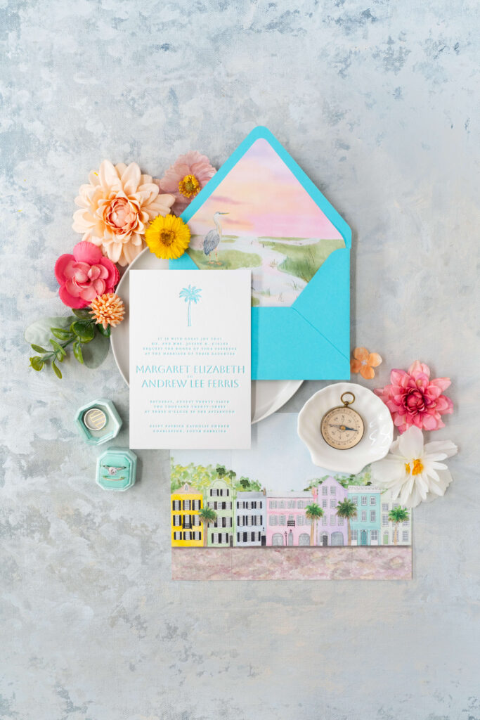 Flatlay of letterpress invitation with watercolor illustrations of Charleston, South Carolina imagery, especially a watercolor gatefold card of Rainbow Row.