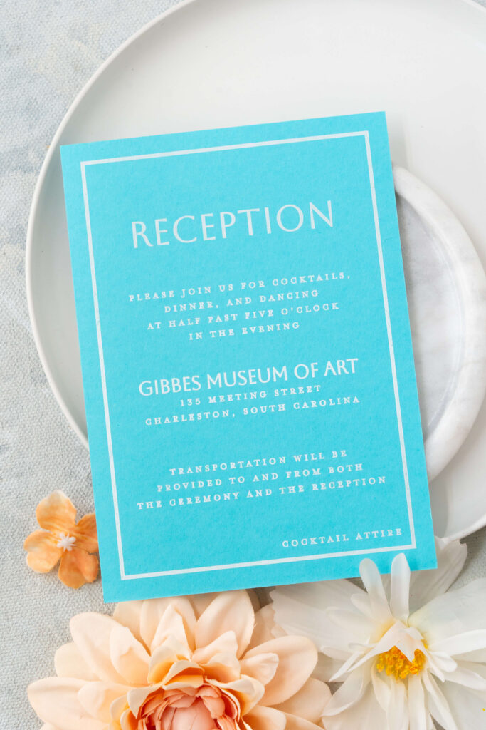 Reception card on turquoise paper with white ink printing detailing the location of the wedding reception at the Gibbes Museum of Art in Charleston, South Carolina.