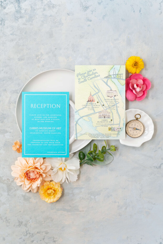 Flatlay of custom wedding invitations, showcasing a Reception Card and a Custom watercolor map of the city of Charleston, South Carolina. Custom map features Rainbow Row, the Gibbes Museum of Art, St. Patrick Catholic Church, Hotel Bennett, and the Arthur Ravenel Bridge.